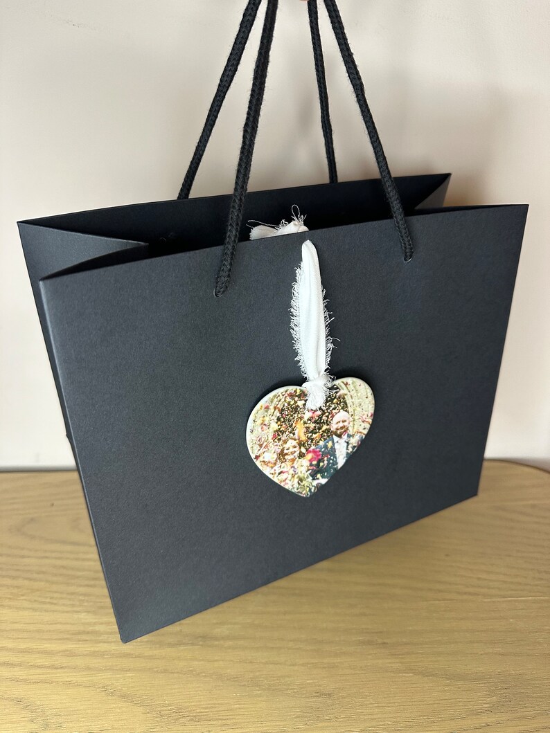 Peut inclure: Un sac cadeau noir avec une &eacute;tiquette en tissu blanc et un ornement photo en forme de c&oelig;ur. La photo montre un groupe de personnes.