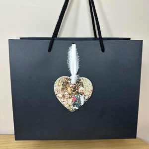 Peut inclure: Un sac cadeau noir avec un ruban blanc et un ornement en forme de c&oelig;ur avec une photo d'un couple et d'invit&eacute;s. L'ornement est suspendu au ruban.