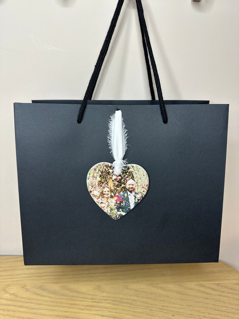Peut inclure: Un sac cadeau noir avec un ruban blanc et un ornement en forme de c&oelig;ur avec une photo d'un couple et d'invit&eacute;s. L'ornement est suspendu au ruban.