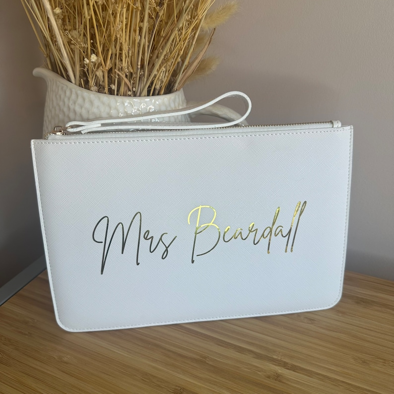 May include: White clutch bag with a gold zipper and a gold inscription that reads "Mrs. Beardall".