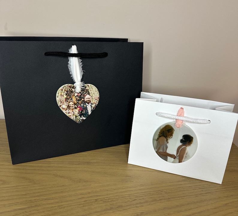 Peut inclure: Deux sacs cadeaux blancs avec des poign&eacute;es en ruban noir et blanc. Un sac a un ornement photo en forme de c&oelig;ur avec une photo de mariage. L'autre sac a un ornement photo rond avec une mari&eacute;e et une demoiselle d'honneur.