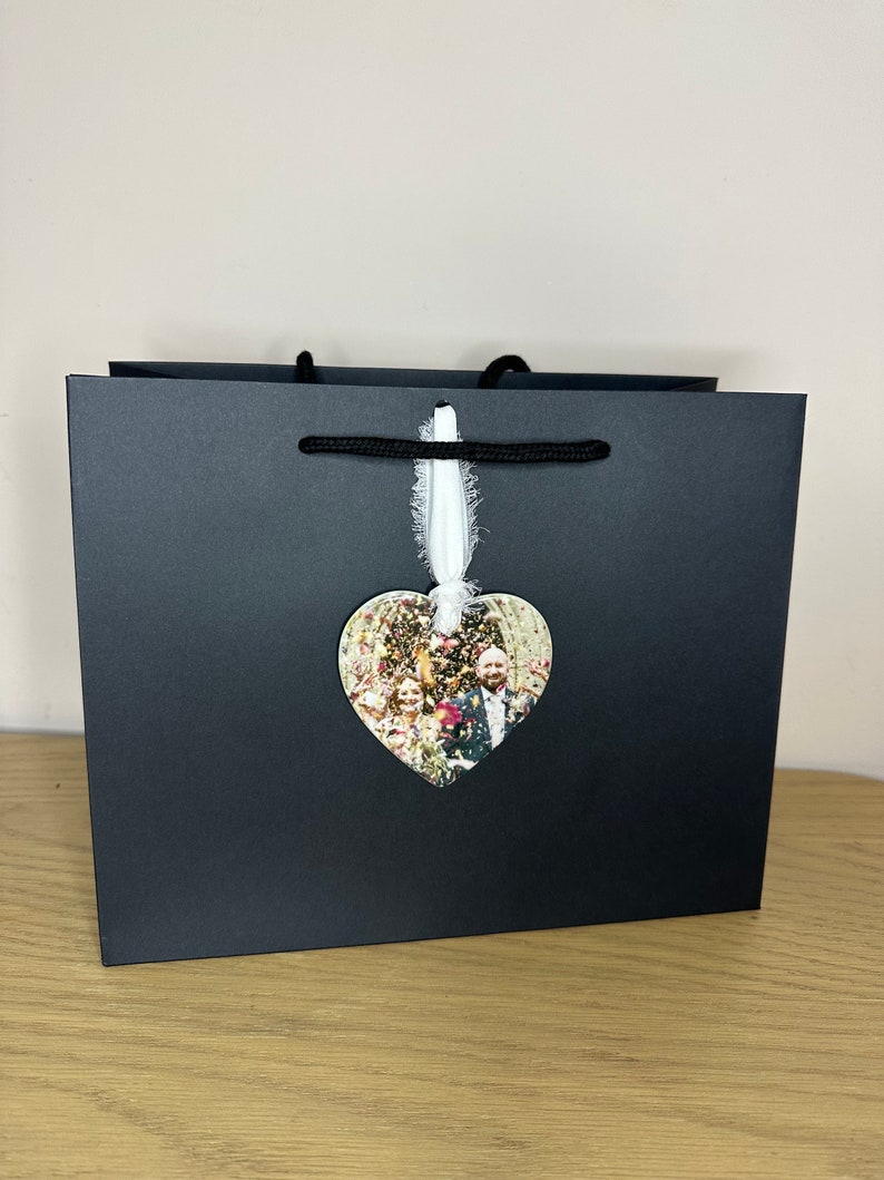Peut inclure: Un sac cadeau noir avec un ruban blanc et un ornement photo en forme de c&oelig;ur repr&eacute;sentant un couple et des invit&eacute;s &agrave; un mariage.