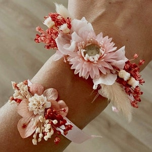 Puede incluir: Dos delicados brazaletes florales con flores secas rosas y rojas en una muñeca. Los brazaletes están hechos con una cinta blanca y presentan una variedad de flores secas, incluyendo margaritas rosas y bayas rojas.