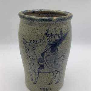 May include: A speckled stoneware jar with a blue illustration of a figure riding a moose, with the year 1991 below. The jar has a cylindrical shape with a slightly flared rim. The illustration is hand-drawn and depicts a whimsical scene.