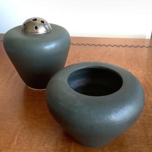 May include: Two dark green ceramic vases. One vase has a silver metal flower frog lid. The other vase is open at the top. Both vases are on a wooden surface. The vases have a smooth, matte finish.