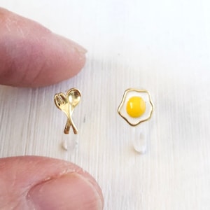 May include: A pair of gold-tone earrings featuring a fried egg and a fork and spoon design.