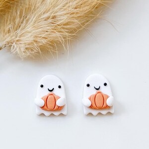 May include: A pair of white ghost-shaped earrings, each holding an orange pumpkin. The ghosts have black eyes and a curved smile. The earrings are set against a white background with dried, beige ornamental grass.
