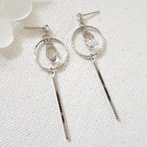 May include: A pair of silver earrings. Each earring has a textured hoop with a dangling crystal and a long, rectangular bar. The earrings are presented on a white, textured surface.
