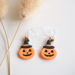 May include: A pair of Halloween-themed clip-on earrings. Each earring features an orange pumpkin with a carved face and a black witch's hat. The earrings are set against a white background.