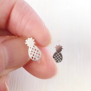 Op de afbeelding: Zilverkleurige ananas oorknopjes. De oorbellen zijn klein, met een getextureerd oppervlak en een gedetailleerd ontwerp. Eén oorbel is scherp, vastgehouden door de vingers van een persoon, terwijl de andere licht wazig is.