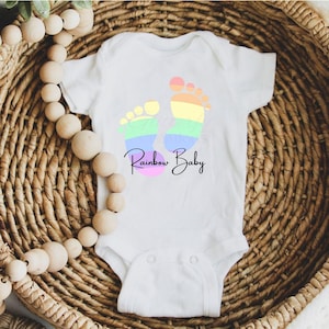 Peut inclure: Combinaison blanche pour bébé avec un motif d'empreinte de pied arc-en-ciel et le texte "Rainbow Baby".