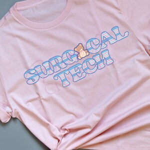 May include: A light pink t-shirt with the words "SURGICAL TECH" in blue and white striped letters. A small teddy bear graphic is above the word "TECH". The t-shirt is knotted at the bottom.