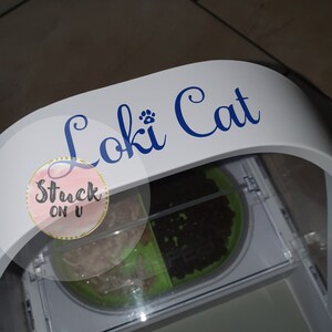May include: White automatic pet feeder with a clear lid and two compartments. The feeder has a blue and white sticker that says "Loki Cat" with a paw print symbol.
