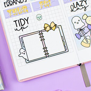 May include: A pink and purple planner with a grid pattern and various Halloween-themed stickers. The stickers include a ghost, a pumpkin, candy, and a notebook. The text on the stickers reads "run errands", "Thursday", "Friday", "Saturday", "lazy day", "grocery run", "tidy up".