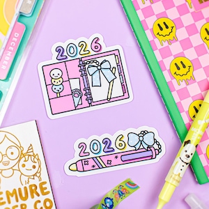 May include: A collection of stationery items on a purple surface. Includes stickers with "2026" and planner designs, a yellow highlighter, a notebook with a pink checkered pattern and smiley faces, and decorative tape. The Demure Paper Co. logo is also visible.