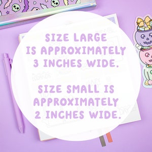 以下が含まれることがあります： 白い円の中に「SIZE LARGE IS APPROXIMATELY 3 INCHES WIDE. SIZE SMALL IS APPROXIMATELY 2 INCHES WIDE.」というテキストが書かれているクローズアップ。円は、ピンクの表面に置かれたノートと、ハロウィーンをテーマにしたキャンディーバケツのステッカーの上に置かれている。