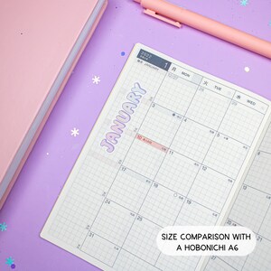May include: A close-up of a small, light pink planner with a gridded layout. The planner is open to January 2022 and features a sticker that says "January" in a colorful, bubbly font. The planner is sitting on a light purple surface with white snowflakes.