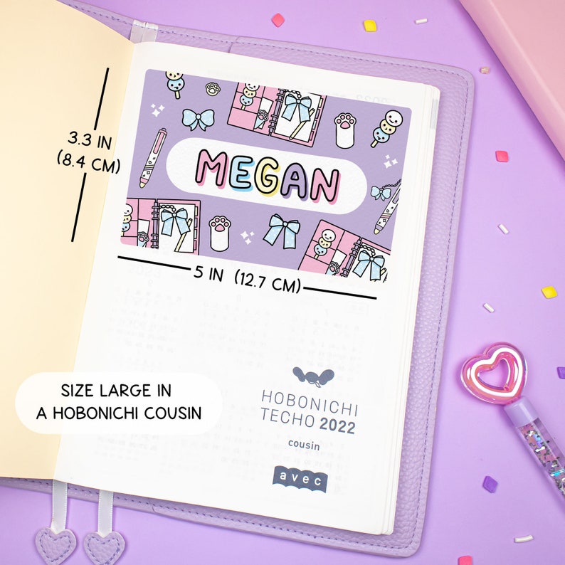 K&ouml;nnte beinhalten: Ein lilafarbener Planer mit einem rosa-wei&szlig;en Aufkleber, auf dem "MEGAN" steht und der niedliche Illustrationen von Schleifen, Pfotenabdr&uuml;cken und japanischen Snacks zeigt. Der Planer ist 12,7 cm breit und 8,4 cm hoch. Auf der Vorderseite steht "Size Large in a Hobonichi Cousin" und "Hobonichi Techo 2022 cousin avec".