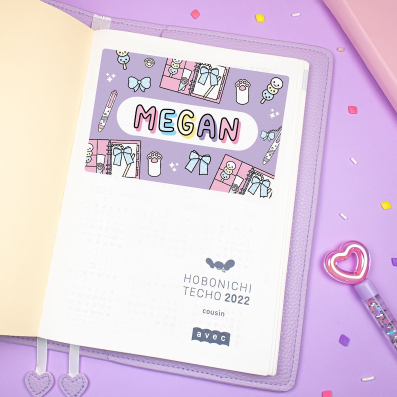 K&ouml;nnte beinhalten: Ein lilafarbener Planer mit einem wei&szlig;en Cover und einem Aufkleber, auf dem "MEGAN" steht. Der Aufkleber hat einen rosa und blauen Hintergrund mit niedlichen Illustrationen von Schleifen, Pfotenabdr&uuml;cken und anderen Designs. Der Planer ist mit "HOBONICHI TECHO 2022 cousin avec" beschriftet.