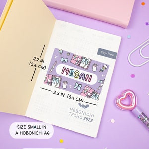 K&ouml;nnte beinhalten: Ein violetter Planer-Aufkleber mit einem bunten Namensschild, auf dem "MEGAN" steht, und niedlichen Illustrationen von Schleifen, Schreibwaren und anderen Kawaii-Gegenst&auml;nden. Der Aufkleber ist 8,4 cm breit und 5,6 cm hoch. Oben und unten steht "Tag frei" und "HOBONICHI TECHNO 2022".