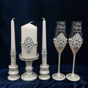 May include: A wedding unity candle set with two taper candles and a larger pillar candle, all with silver embellishments. Two champagne flutes with silver embellishments and the text "Mr. Alford 08/26/2023" and "Mrs. Alford 08/26/2023" are also included.
