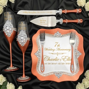 Puede incluir: Un juego de servir pastel para el séptimo aniversario de bodas con un plato de color cobre, dos copas de champán y un cuchillo y servidor para pastel. El plato tiene un centro blanco con letras doradas que dicen "7º aniversario de boda Charlie & Ella 12/22/2014-12/22/2021". Las copas y las piezas de servicio están decoradas con diamantes de imitación.