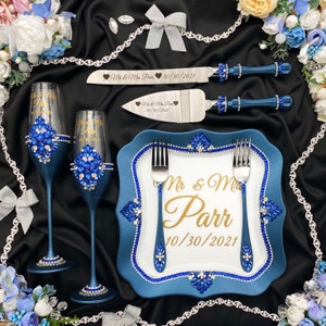 May include: Blue wedding cake serving set featuring a blue and white plate, two blue champagne flutes, and two blue and silver cake knives. The plate and flutes are decorated with rhinestones. The knives are engraved with "Mr. & Mrs. Parr" and the date "10/30/2021".