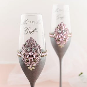 Può includere: Due flûte da champagne con steli e basi argentate. I bicchieri sono decorati con cristalli rosa e trasparenti. Il testo "Mr & Mrs Griffin" è inciso su un bicchiere. Tema matrimonio.