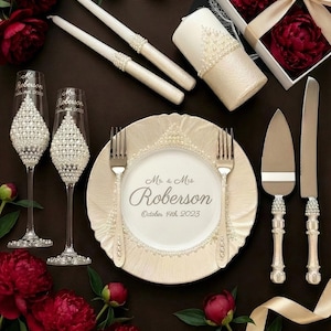 Puede incluir: Elegante mesa de boda con copas de champán, plato, tenedores, servidor de pastel y velas, todos adornados con perlas. El plato está inscrito con "Mr. & Mrs. Roberson October 14th, 2023". Peonías borgoña y una caja de regalo con una cinta completan el arreglo.