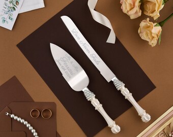 May include: A set of two white cake knives with pearl-like accents. The larger knife has the text "Mr. & Mrs.  Ferraro June 1st, 2024" engraved on the blade. The smaller knife has the text "Love Never Fails" engraved on the blade.