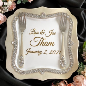 May include: A white ceramic cake plate with a gold rim and a diamond-like design. The plate has two forks and the text "Lisa & Joe Thom January 2, 2021" in gold lettering.