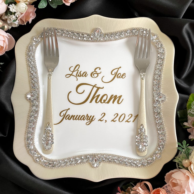 May include: A white ceramic cake plate with a gold rim and a diamond-like design. The plate has two forks and the text "Lisa & Joe Thom January 2, 2021" in gold lettering.