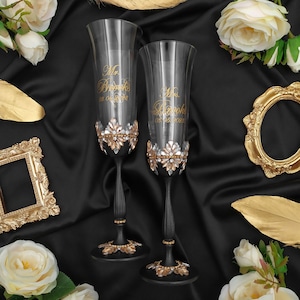 May include: Two black champagne flutes with gold lettering and rhinestones. The flutes are decorated with gold and silver embellishments. The text on the flutes reads "Mr. Brooks 08.15.2023" and "Mrs. Brooks 08.15.2023".