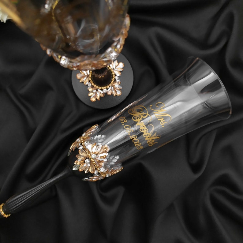 May include: Two champagne flutes with gold detailing and rhinestones. The flute on the right has the text "Mrs. Rosales 05.05.2023" engraved on it.