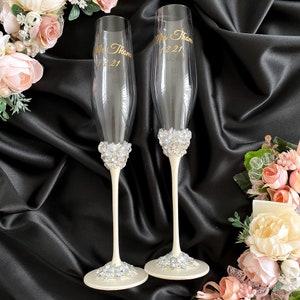 May include: Two champagne flutes with white stems and bases decorated with rhinestones. The flutes are personalized with the text "Mr. Thom 12.21" and "Mrs. Thom 12.21" in gold lettering.