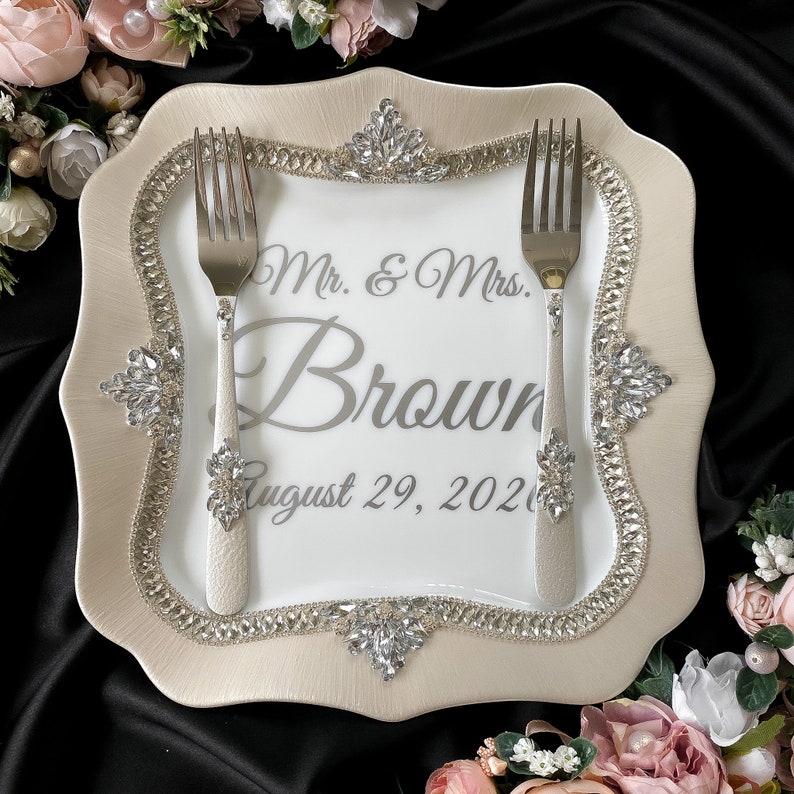 Puede incluir: Un plato de pastel de cer&aacute;mica blanca con un borde decorativo de diamantes de imitaci&oacute;n. El plato tiene el texto "Mr. & Mrs. Brown August 29, 2021" impreso en &eacute;l. Dos tenedores plateados se colocan en el plato.