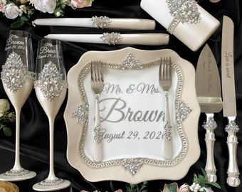 May include: A wedding cake serving set with a white and silver design. The set includes a cake knife, two forks, two champagne flutes, and a cake plate. The cake plate has a white background with the text "Mr. & Mrs. Brown August 29, 2020" in silver lettering. The champagne flutes and cake knife have a silver band with rhinestones.