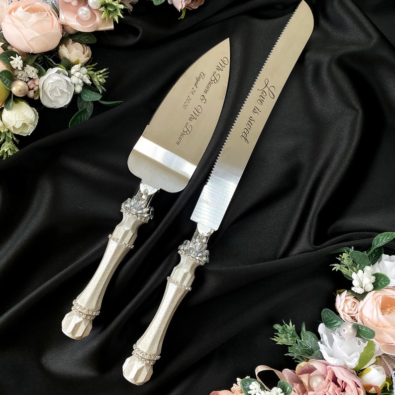 Puede incluir: Un juego de dos cuchillos para pastel de bodas plateados con mangos blancos decorados con diamantes de imitaci&oacute;n. Los cuchillos est&aacute;n grabados con el texto "Jane & Matt" y "Mr & Mrs Dalton October 24, 2020".