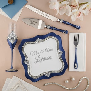 Puede incluir: Un plato de pastel azul y blanco con fondo blanco y el texto "Mis 15 Años Larissa" en letras plateadas. El plato está decorado con pedrería alrededor del borde. Hay un cuchillo para pastel azul y plateado, un servidor para pastel azul y plateado, una flauta de champán azul y plateada, un tenedor azul y plateado, una servilleta blanca, una cadena de perlas y dos aretes de perlas.