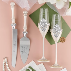 Puede incluir: Un juego de cuchillo y servidor para cortar pastel de bodas, ambos con mangos blancos y cuchillas plateadas. El servidor está grabado con "Mr. & Mrs. Grimaldi June 2, 2023". Dos copas de champán con bases blancas y tallos plateados están decoradas con diamantes de imitación.