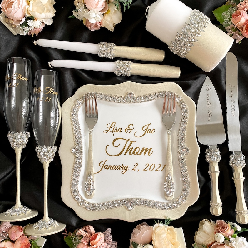 May include: A wedding cake cutting set with a white cake knife and server with crystal-embellished handles. The set is displayed on a white plate with a gold rim and a gold inscription that reads "Lisa & Joe Thom January 2, 2021". Two champagne flutes with crystal embellishments are also included.