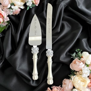 May include: A set of two white cake cutting knives with silver blades. The handles are decorated with rhinestones. The larger knife is engraved with "January 2, 2021" and the smaller knife is engraved with "Mr. & Mrs. Them."