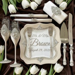 Könnte beinhalten: Hochzeitstisch mit Sektgläsern, einem Teller, Gabeln, Kuchenmesser und -servierer sowie Kerzen. Auf dem Teller steht "Mr. & Mrs. Brown August 29, 2020". Die Gläser und der Teller sind mit Strass verziert. Weiße Tulpen sind um die Artikel angeordnet.