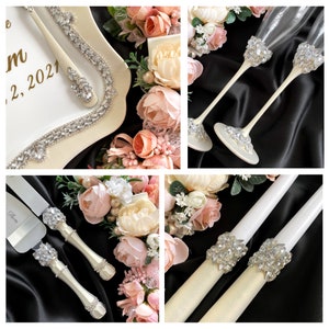 May include: Wedding cake cutting set with two ivory handles and silver blades, two ivory candles with crystal embellishments, and two crystal-embellished champagne flutes. The set is displayed on a black background with pink and white flowers.