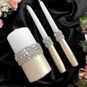 May include: Three white wedding candles with a decorative band of rhinestones. The candles are on a black satin fabric background.