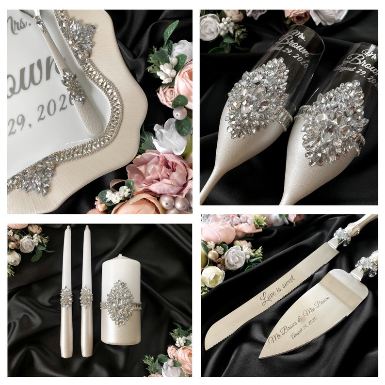 Puede incluir: Un juego de corte de pastel de boda con un cuchillo y un servidor de pastel blancos, dos velas blancas y dos copas de champ&aacute;n. El cuchillo y el servidor tienen mangos plateados y est&aacute;n decorados con diamantes de imitaci&oacute;n. Las copas de champ&aacute;n tambi&eacute;n est&aacute;n decoradas con diamantes de imitaci&oacute;n y tienen el texto "Mr. Brown & Mrs. Brown August 29, 2020" grabado en ellas. Las velas tambi&eacute;n est&aacute;n decoradas con diamantes de imitaci&oacute;n. El juego est&aacute; sobre un fondo negro.