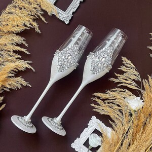 Puede incluir: Dos copas de champán blancas con purpurina plateada y pedrería. Las copas están decoradas con las palabras "Mr." y "Mrs." en cursiva.