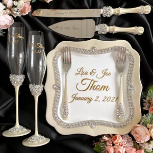 May include: A white cake plate with a gold border and rhinestones. The plate has a white background with the text "Lisa & Joe Thom January 2, 2021" in gold lettering. Two silver forks are placed on the plate. Two champagne flutes with rhinestones are on the left side of the plate. The plate is surrounded by pink flowers.
