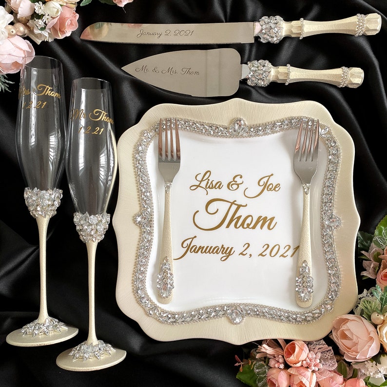 May include: A white cake plate with a gold border and rhinestones. The plate has a white background with the text "Lisa & Joe Thom January 2, 2021" in gold lettering. Two silver forks are placed on the plate. Two champagne flutes with rhinestones are on the left side of the plate. The plate is surrounded by pink flowers.