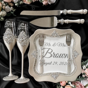 Puede incluir: Un juego de servir pastel de bodas con un plato de pastel blanco, dos tenedores y un cuchillo. El plato tiene un borde decorativo de diamantes de imitaci&oacute;n y el texto "Mr. & Mrs. Brown August 29, 2020". Tambi&eacute;n se incluyen dos copas de champ&aacute;n con diamantes de imitaci&oacute;n. El cuchillo y la esp&aacute;tula de servir tienen mangos plateados con diamantes de imitaci&oacute;n.